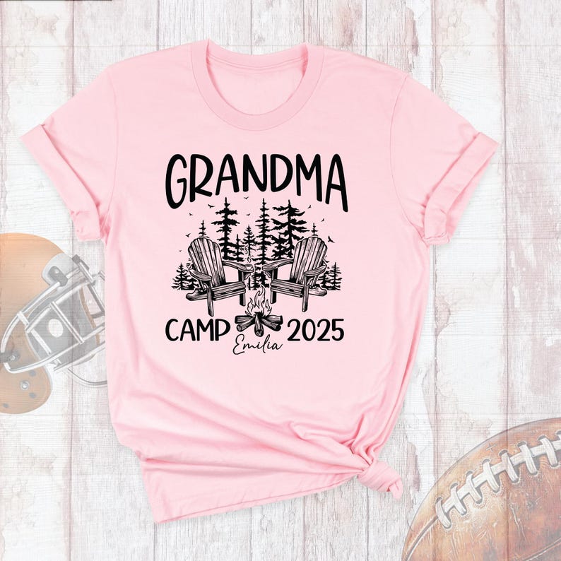 May include: A light pink t-shirt with the words "GRANDMA CAMP 2025" in black. The design includes two wooden chairs, a campfire, and trees. The name "Emilia" is printed below the campfire.