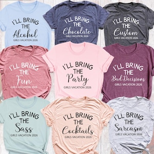May include: A collection of nine short-sleeved crew neck t-shirts in various colors including light blue, dark gray, pink, and mauve. Each shirt features the text I'LL BRING THE followed by a unique word and GIRLS VACATION 2026.