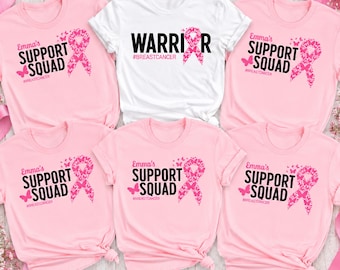 May include: Five short-sleeved t-shirts arranged in a group. Four shirts are light pink with the black text "Emma's SUPPORT SQUAD" and a pink butterfly-patterned ribbon. One white shirt features the black text "WARRIOR" with a pink ribbon.