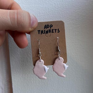 May include: A pair of pink teapot-shaped dangle earrings with white accents. The earrings are on a brown card with the text "ADP TRINKETS".