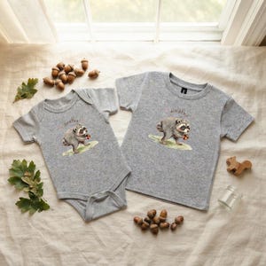 May include: A heather grey short-sleeve bodysuit and a matching heather grey short-sleeve t-shirt, both featuring a watercolour illustration of a raccoon holding an acorn with the text "Jimothy" printed in a cursive script above the animal.
