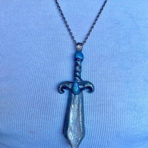 Peut inclure: Un collier pendentif en forme d'épée argentée avec une lame texturée aux reflets irisés, une poignée torsadée et une perle sphérique bleu vif au sommet. La garde de l'épée est ornée d'une petite pierre bleue en forme de goutte d'eau.