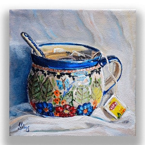 May include: A colourful ceramic teacup with a floral and butterfly design, filled with tea and a tea bag hanging from the rim. A spoon rests on the rim of the cup. The cup is sitting on a white cloth.