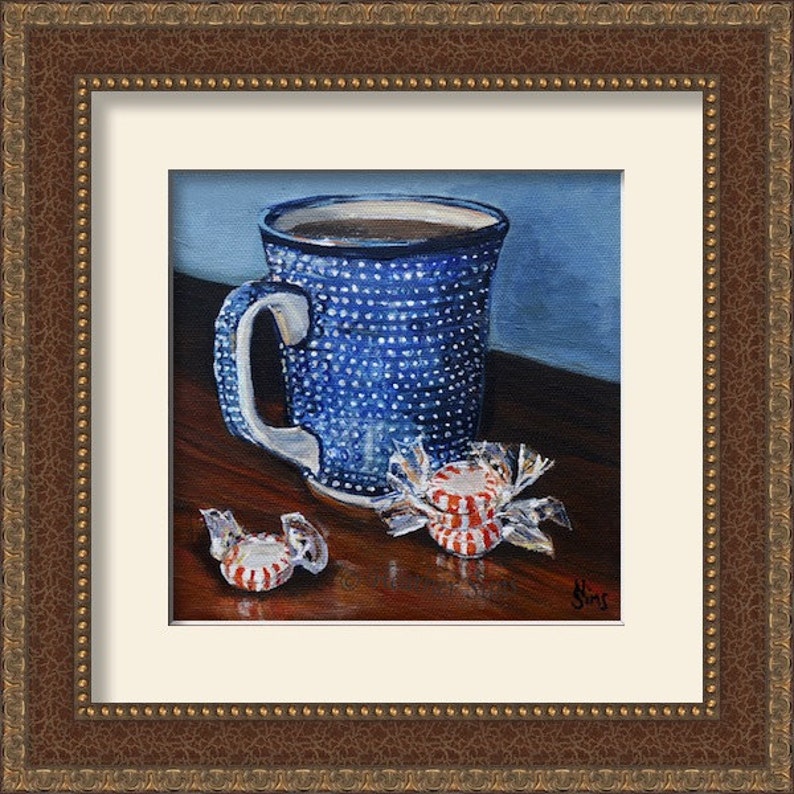 Puede incluir: Una taza de cer&aacute;mica azul y blanca con asa marr&oacute;n llena de caf&eacute;, sentada sobre una mesa de madera con tres caramelos de menta roja y blanca sin envolver.