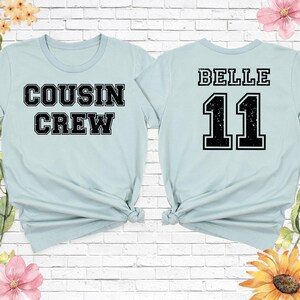 Op de afbeelding: Twee lichtblauwe t-shirts. E&eacute;n shirt heeft de tekst "COUSIN CREW" in zwarte blokletters. Het andere shirt heeft de naam "BELLE" boven het nummer "11" in zwarte blokletters. Beide shirts zijn aan de onderkant geknoopt.