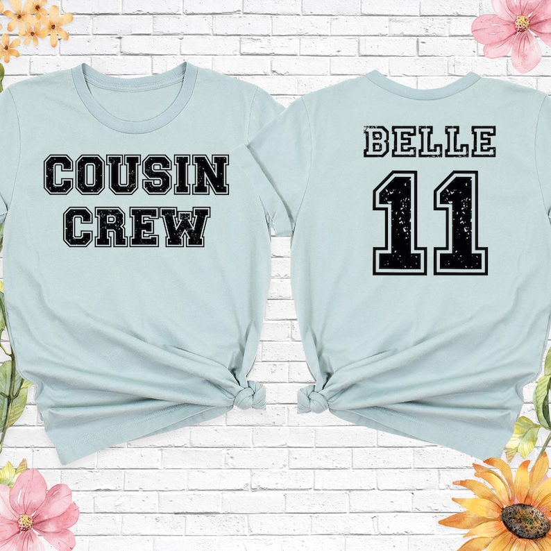 Op de afbeelding: Twee lichtblauwe t-shirts. E&eacute;n shirt heeft de tekst "COUSIN CREW" in zwarte blokletters. Het andere shirt heeft de naam "BELLE" boven het nummer "11" in zwarte blokletters. Beide shirts zijn aan de onderkant geknoopt.