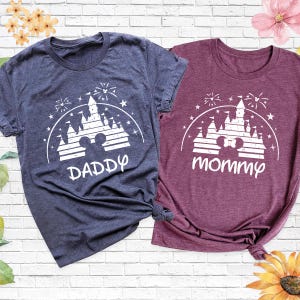 May include: Two short-sleeved t-shirts, one navy blue and one burgundy, with a white graphic of a castle, fireworks, and the text "DADDY" and "mommy". The shirts are knotted at the bottom.