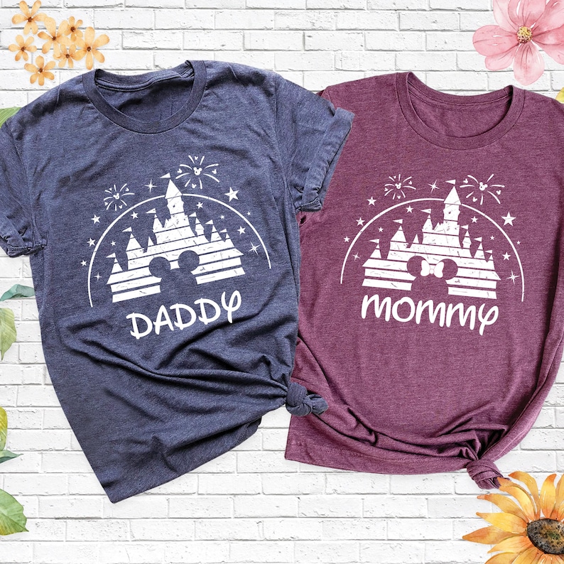 May include: Two short-sleeved t-shirts, one navy blue and one burgundy, with a white graphic of a castle, fireworks, and the text "DADDY" and "mommy". The shirts are knotted at the bottom.