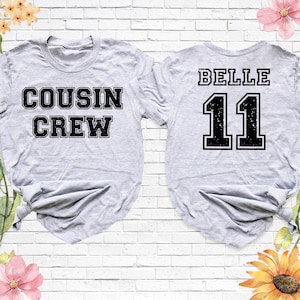 Op de afbeelding: Twee lichtgrijze t-shirts. E&eacute;n shirt heeft de woorden "COUSIN CREW" in een zwarte, blokletter. Het andere shirt heeft het woord "BELLE" boven het nummer "11" in een zwarte, blokletter. De shirts staan op een witte bakstenen achtergrond.