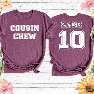 Op de afbeelding: Twee bordeauxrode t-shirts. E&eacute;n shirt heeft "COUSIN CREW" in witte blokletters. Het andere shirt heeft "ZANE" boven het nummer "10" in witte blokletters. De shirts worden gepresenteerd op een witte bakstenen muur met bloemaccenten.