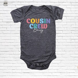 Op de afbeelding: Een donkergrijze babybody met de tekst "COUSIN CREW" in kleurrijke letters en de naam "Emily" in witte letters. De body heeft korte mouwen en drie drukkers aan de onderkant.