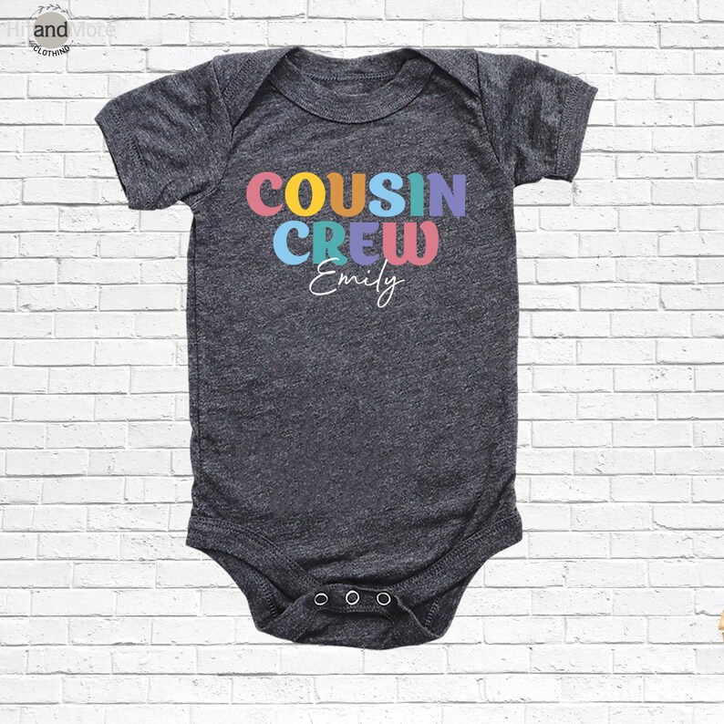 Op de afbeelding: Een donkergrijze babybody met de tekst "COUSIN CREW" in kleurrijke letters en de naam "Emily" in witte letters. De body heeft korte mouwen en drie drukkers aan de onderkant.