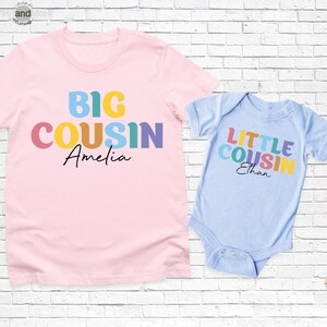 Op de afbeelding: Een roze t-shirt met de woorden "BIG COUSIN Amelia" in kleurrijke letters. Daarnaast een lichtblauwe babyromper met "LITTLE COUSIN Ethan" in vergelijkbare kleuren. Beide items worden gepresenteerd tegen een witte bakstenen muur.