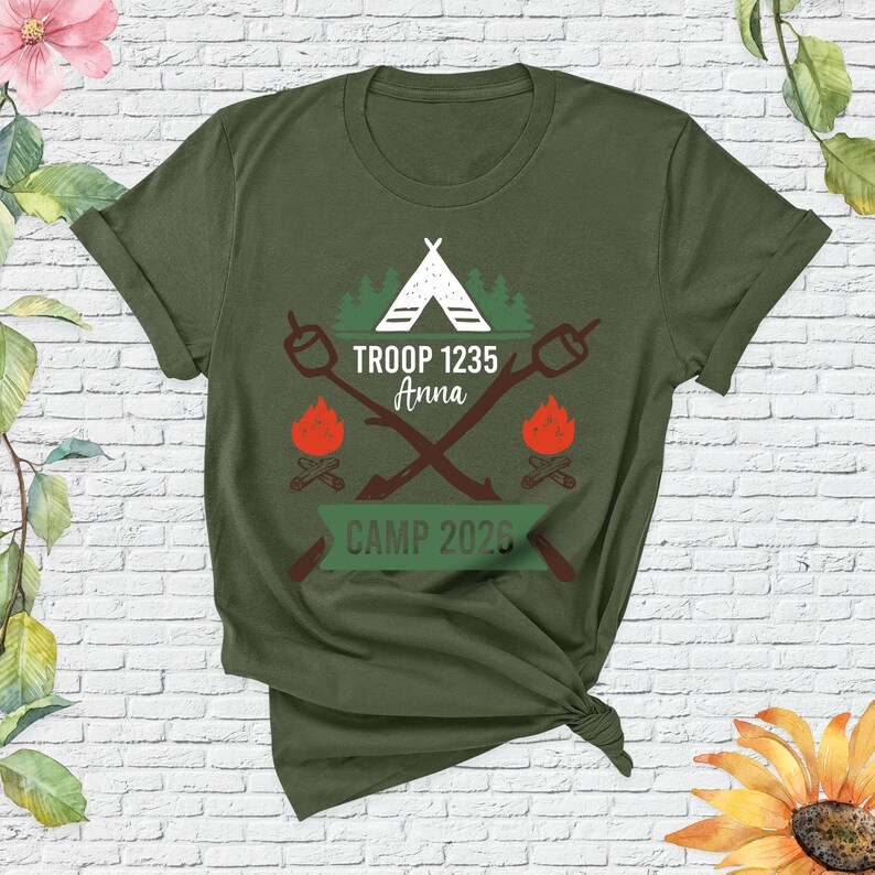 May include: Olive green t-shirt with a camping-themed graphic. The design includes a white tent, crossed marshmallow roasting sticks, campfires, and the text "TROOP 1235 Anna" and "CAMP 2026".