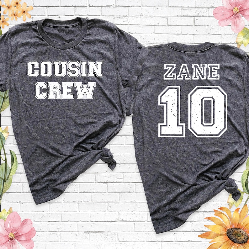 Op de afbeelding: Twee donkergrijze t-shirts. E&eacute;n shirt heeft "COUSIN CREW" in witte blokletters. Het andere shirt heeft "ZANE" en het nummer "10" in witte blokletters. Beide shirts zijn aan de onderkant geknoopt.