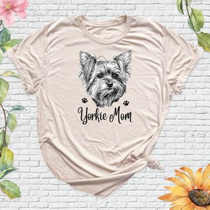 May include: A beige t-shirt with a black and white illustration of a Yorkshire Terrier and the text "Yorkie Mom".
