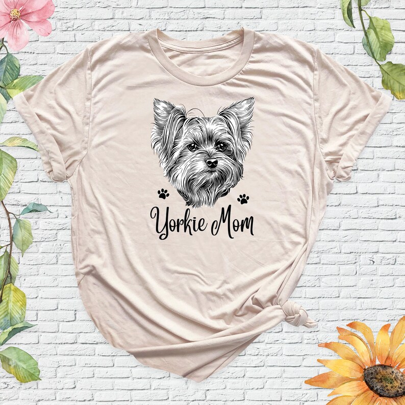 May include: A beige t-shirt with a black and white illustration of a Yorkshire Terrier and the text "Yorkie Mom".