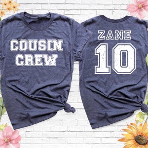 Op de afbeelding: Twee marineblauwe t-shirts. E&eacute;n shirt heeft de woorden "COUSIN CREW" in witte blokletters. Het andere shirt heeft de naam "ZANE" en het nummer "10" in witte blokletters. Beide shirts staan op een witte bakstenen achtergrond.