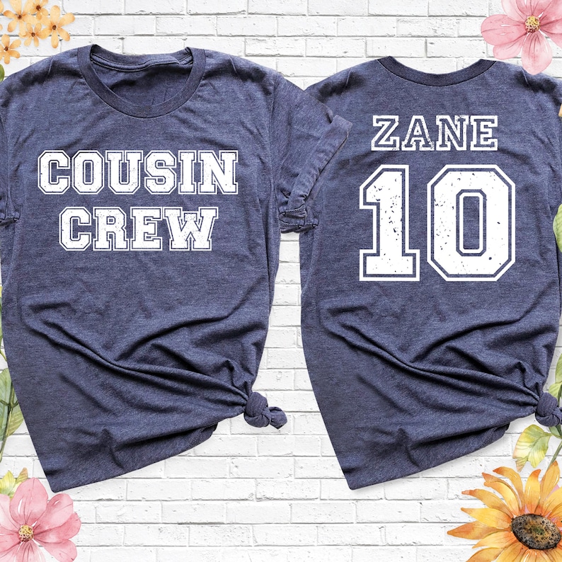 Op de afbeelding: Twee marineblauwe t-shirts. E&eacute;n shirt heeft de woorden "COUSIN CREW" in witte blokletters. Het andere shirt heeft de naam "ZANE" en het nummer "10" in witte blokletters. Beide shirts staan op een witte bakstenen achtergrond.
