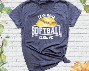 May include: A heather navy blue t-shirt featuring a yellow softball graphic with white text that reads TEAM NAME, SOFTBALL, and CLARA #17. The shirt has short sleeves, a crew neckline, and a knotted hem detail on the lower right side.