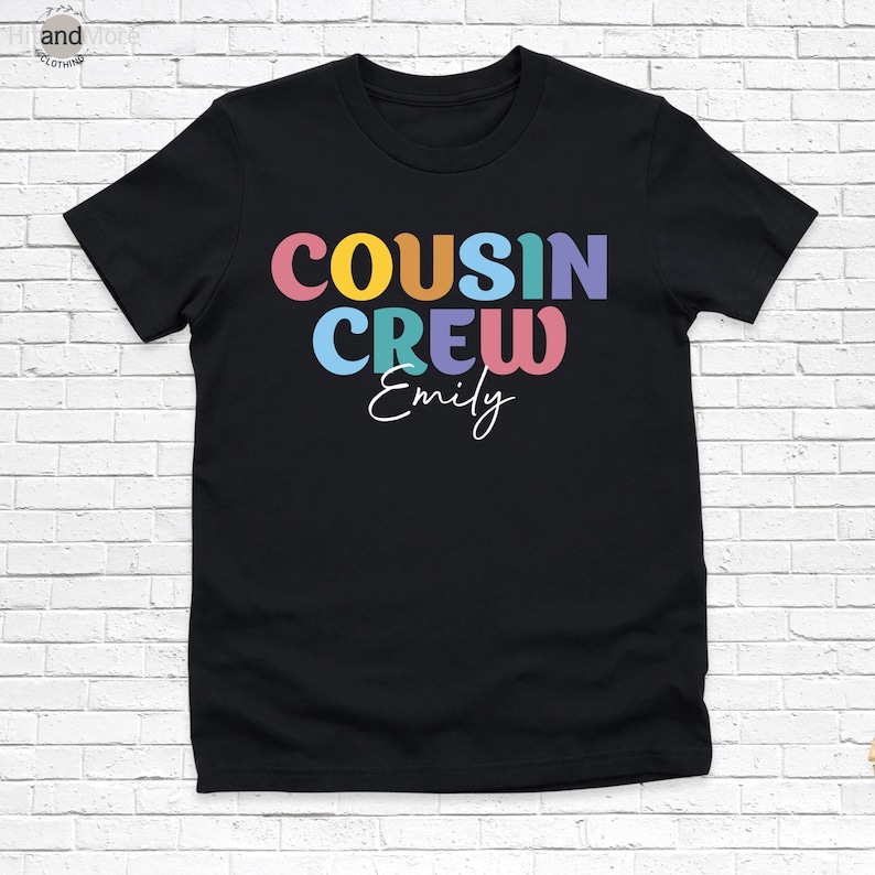 Op de afbeelding: Zwart T-shirt met de tekst "COUSIN CREW" in kleurrijke, retro letters. Onder de tekst staat de naam "Emily" in een sierlijk lettertype. Het shirt heeft een ronde hals.