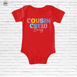 Op de afbeelding: Een rode babybody met de tekst "COUSIN CREW Emily" in kleurrijke letters. De tekst is in een speels lettertype, geschikt voor baby's en peuters. De body heeft korte mouwen en drukknoopjes aan de onderkant.