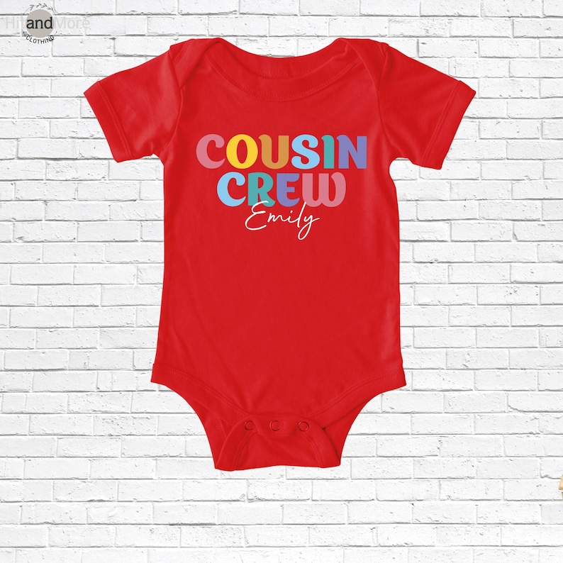 Op de afbeelding: Een rode babybody met de tekst "COUSIN CREW Emily" in kleurrijke letters. De tekst is in een speels lettertype, geschikt voor baby's en peuters. De body heeft korte mouwen en drukknoopjes aan de onderkant.
