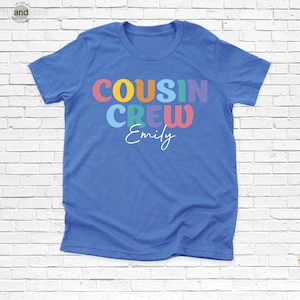 Op de afbeelding: Een blauw t-shirt met de woorden "COUSIN CREW" in kleurrijke letters. Onder de woorden staat de naam "Emily" in een sierlijk lettertype. Het shirt is gemaakt van een zacht materiaal.