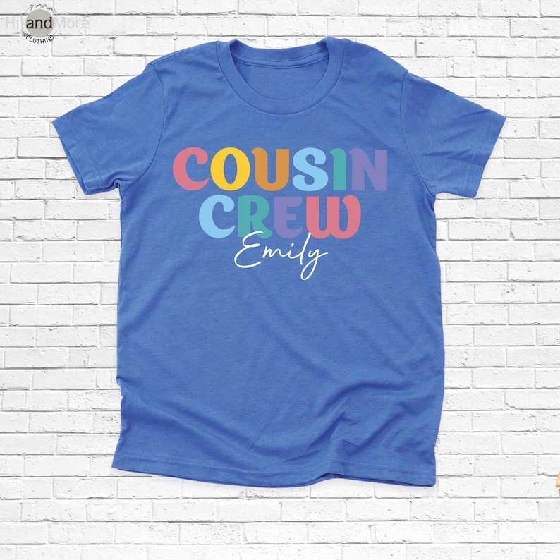 Op de afbeelding: Een blauw t-shirt met de woorden "COUSIN CREW" in kleurrijke letters. Onder de woorden staat de naam "Emily" in een sierlijk lettertype. Het shirt is gemaakt van een zacht materiaal.