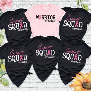 May include: Five black t-shirts with pink ribbon designs. The shirts say "Support Squad" with a pink ribbon and the hashtag "#TEAMNAME". One pink t-shirt says "Warrior" with a pink ribbon and the hashtag "#TEAMNAME".