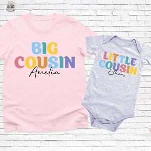 Op de afbeelding: Een roze t-shirt met de woorden "BIG COUSIN Amelia" in kleurrijke blokletters. Daarnaast een grijze babyromper met "LITTLE COUSIN Ethan" in vergelijkbare kleuren. Beide items staan op een witte bakstenen achtergrond.