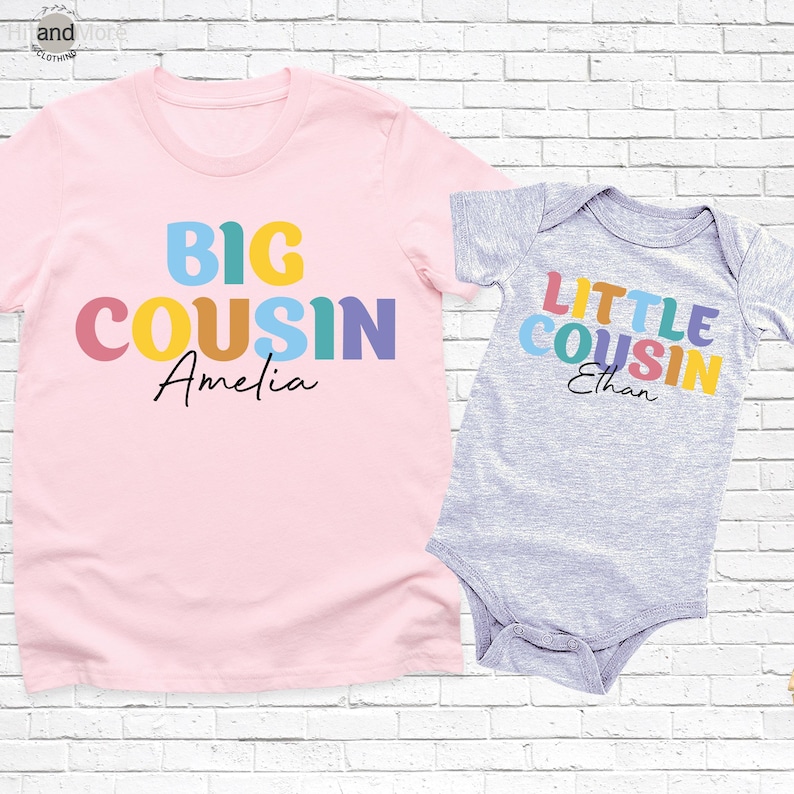 Op de afbeelding: Een roze t-shirt met de woorden "BIG COUSIN Amelia" in kleurrijke blokletters. Daarnaast een grijze babyromper met "LITTLE COUSIN Ethan" in vergelijkbare kleuren. Beide items staan op een witte bakstenen achtergrond.