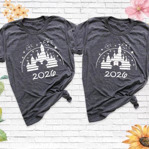 May include: Two dark gray t-shirts with a white graphic featuring a castle, fireworks, and the year "2026". The shirts are knotted at the bottom. The background is a white brick wall with floral accents.
