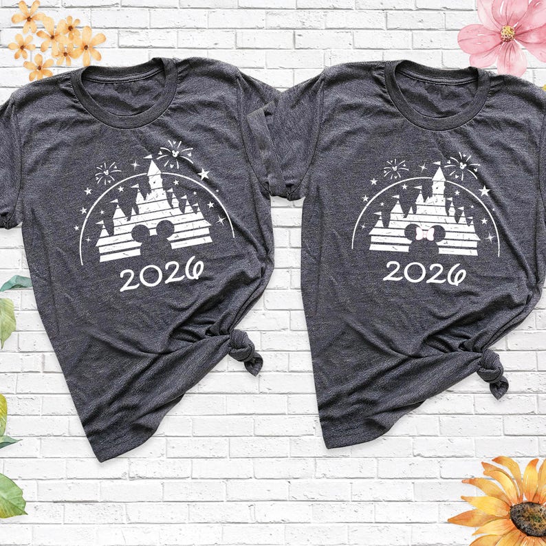 May include: Two dark gray t-shirts with a white graphic featuring a castle, fireworks, and the year "2026". The shirts are knotted at the bottom. The background is a white brick wall with floral accents.