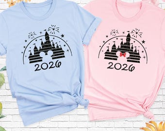 May include: A light blue t-shirt and a light pink t-shirt, each featuring a black graphic of a castle silhouette, fireworks, stars, and the year 2026. The pink shirt includes a small red and white polka dot bow on the castle icon.