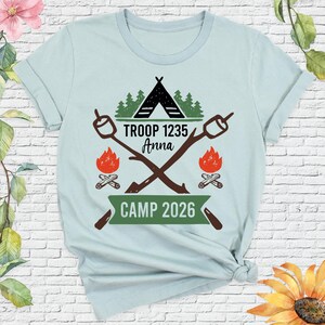 May include: Light blue t-shirt with a camping theme. The design includes a black tent, green trees, crossed marshmallow roasting sticks, and flames. Text reads "TROOP 1235 Anna" and "CAMP 2026".
