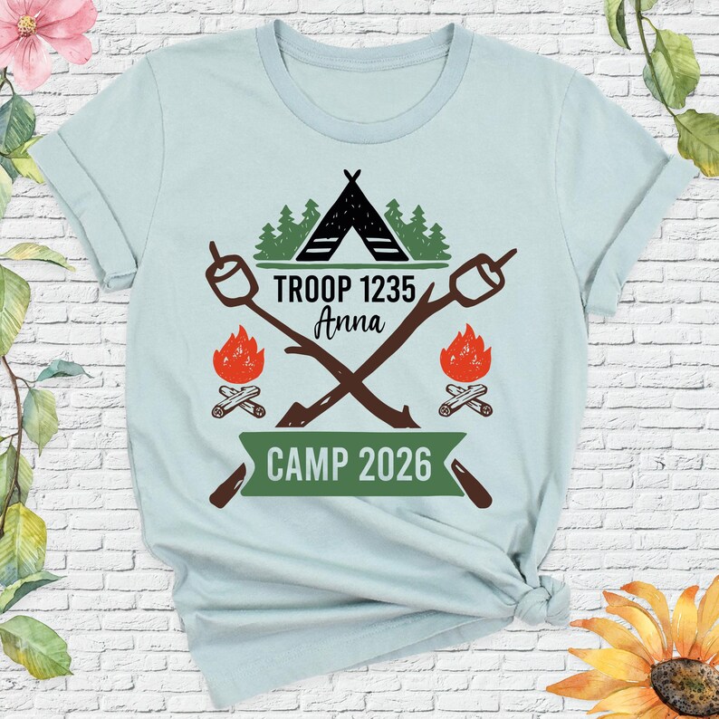 May include: Light blue t-shirt with a camping theme. The design includes a black tent, green trees, crossed marshmallow roasting sticks, and flames. Text reads "TROOP 1235 Anna" and "CAMP 2026".