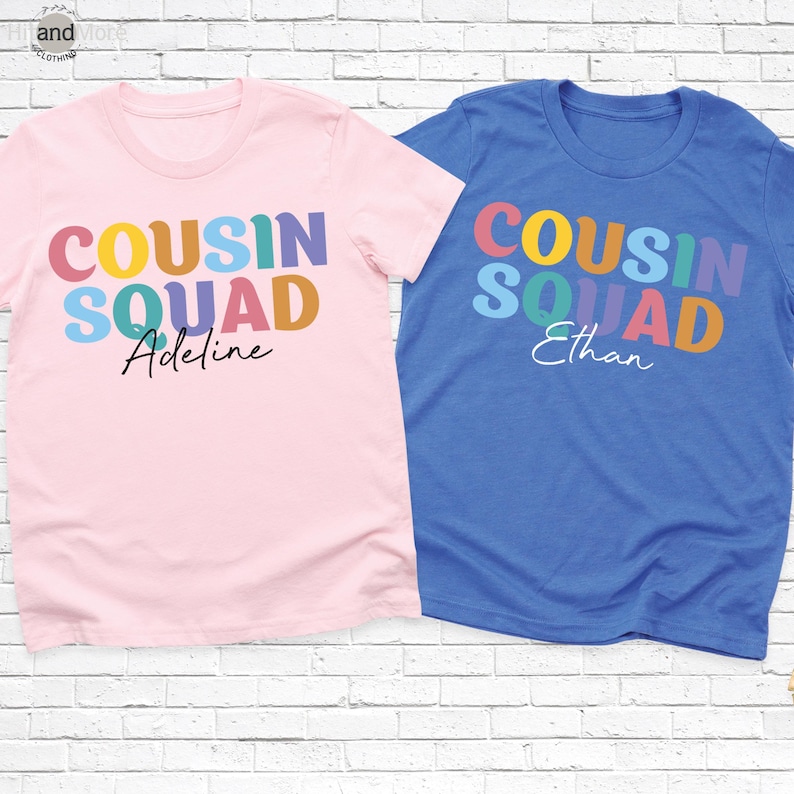 Op de afbeelding: Twee T-shirts met korte mouwen. E&eacute;n is lichtroze met de woorden "COUSIN SQUAD" in kleurrijke blokletters en de naam "Adeline" in cursief. De andere is blauw met "COUSIN SQUAD" en de naam "Ethan".