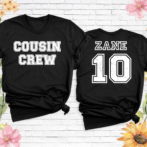 Op de afbeelding: Twee zwarte t-shirts met witte tekst. E&eacute;n shirt leest "COUSIN CREW" en de andere "ZANE 10". De tekst heeft een lettertype in collegestijl. De shirts worden gepresenteerd tegen een witte bakstenen muur met bloemaccenten.