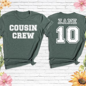 Op de afbeelding: Twee bosgroene t-shirts. E&eacute;n shirt heeft "COUSIN CREW" in witte blokletters. Het andere shirt heeft "ZANE" en het nummer "10" in witte blokletters. Beide shirts hebben een ronde hals en korte mouwen.