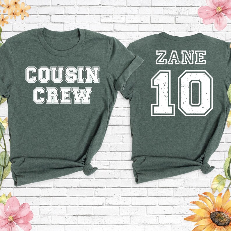 Op de afbeelding: Twee bosgroene t-shirts. E&eacute;n shirt heeft "COUSIN CREW" in witte blokletters. Het andere shirt heeft "ZANE" en het nummer "10" in witte blokletters. Beide shirts hebben een ronde hals en korte mouwen.