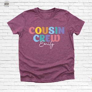 Op de afbeelding: Een bordeauxrood t-shirt met de woorden "COUSIN CREW" in kleurrijke blokletters en de naam "Emily" in een sierlijk lettertype. Het shirt is gemaakt van een zacht materiaal en heeft korte mouwen.