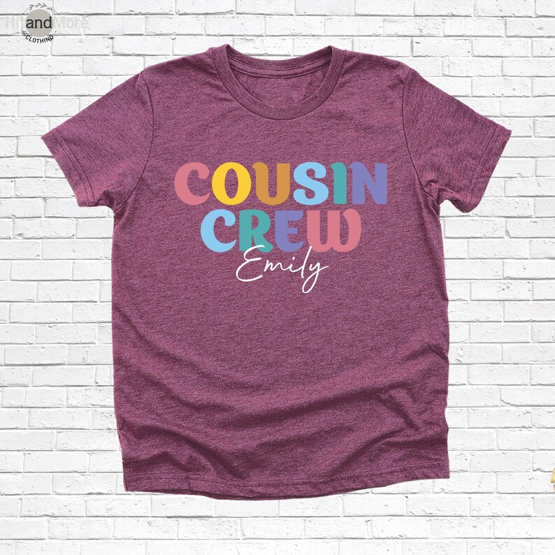 Op de afbeelding: Een bordeauxrood t-shirt met de woorden "COUSIN CREW" in kleurrijke blokletters en de naam "Emily" in een sierlijk lettertype. Het shirt is gemaakt van een zacht materiaal en heeft korte mouwen.