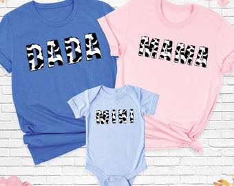 Könnte beinhalten: Ein blaues T-Shirt mit dem Text DADA, ein rosafarbenes T-Shirt mit dem Text MAMA und ein hellblauer Babybody mit dem Text MINI. Jedes Kleidungsstück zeigt den Text in einem schwarz-weißen Kuhfleckenmuster auf der Vorderseite der Artikel.