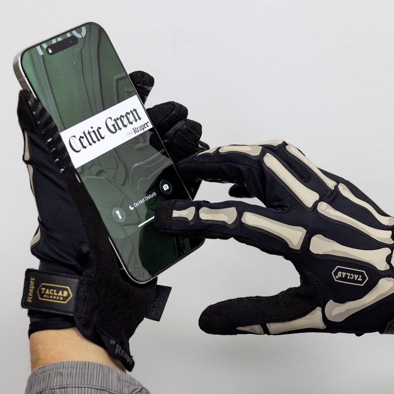May include: Black tactical gloves featuring a white skeleton bone print on the back of the hand and fingers. The gloves have a black wrist strap with a gold and black patch reading TACLAB GLOVES and the word Reaper. A smartphone is being held.