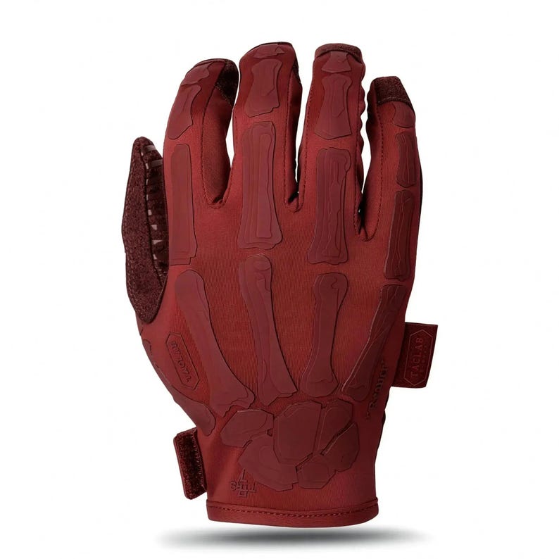May include: A dark red tactical glove featuring a raised, tonal skeleton hand design on the back. The glove includes a hook-and-loop wrist closure, a textured thumb panel, and a small rectangular tag on the side with the text TACLAB printed on it.