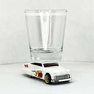 Puede incluir: Un coche de juguete blanco con un diseño de llama roja y amarilla está equilibrado sobre un vaso de chupito de vidrio transparente.