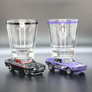 Puede incluir: Dos coches de juguete, uno negro y otro morado, están en equilibrio sobre dos vasos de chupito de vidrio transparente. Los vasos de chupito tienen una banda negra y morada alrededor de la parte superior.