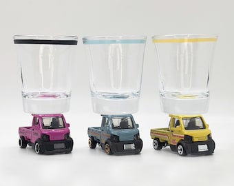Op de afbeelding: Drie speelgoed pick-up trucks, elk met een andere carrosseriekleur, zijn gebalanceerd op heldere glazen shotglazen. De trucks zijn roze, blauw en geel.