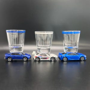 以下が含まれることがあります： 青い縁取りの透明なショットグラス3つが、それぞれミニチュアのミニカーの上に置かれています。ミニカーは、青いセダン、赤と青のレーシングデカールが施された白いスバルラリースタイルのクーペ、そしてリアスポイラー付きの青いスポーツクーペです。